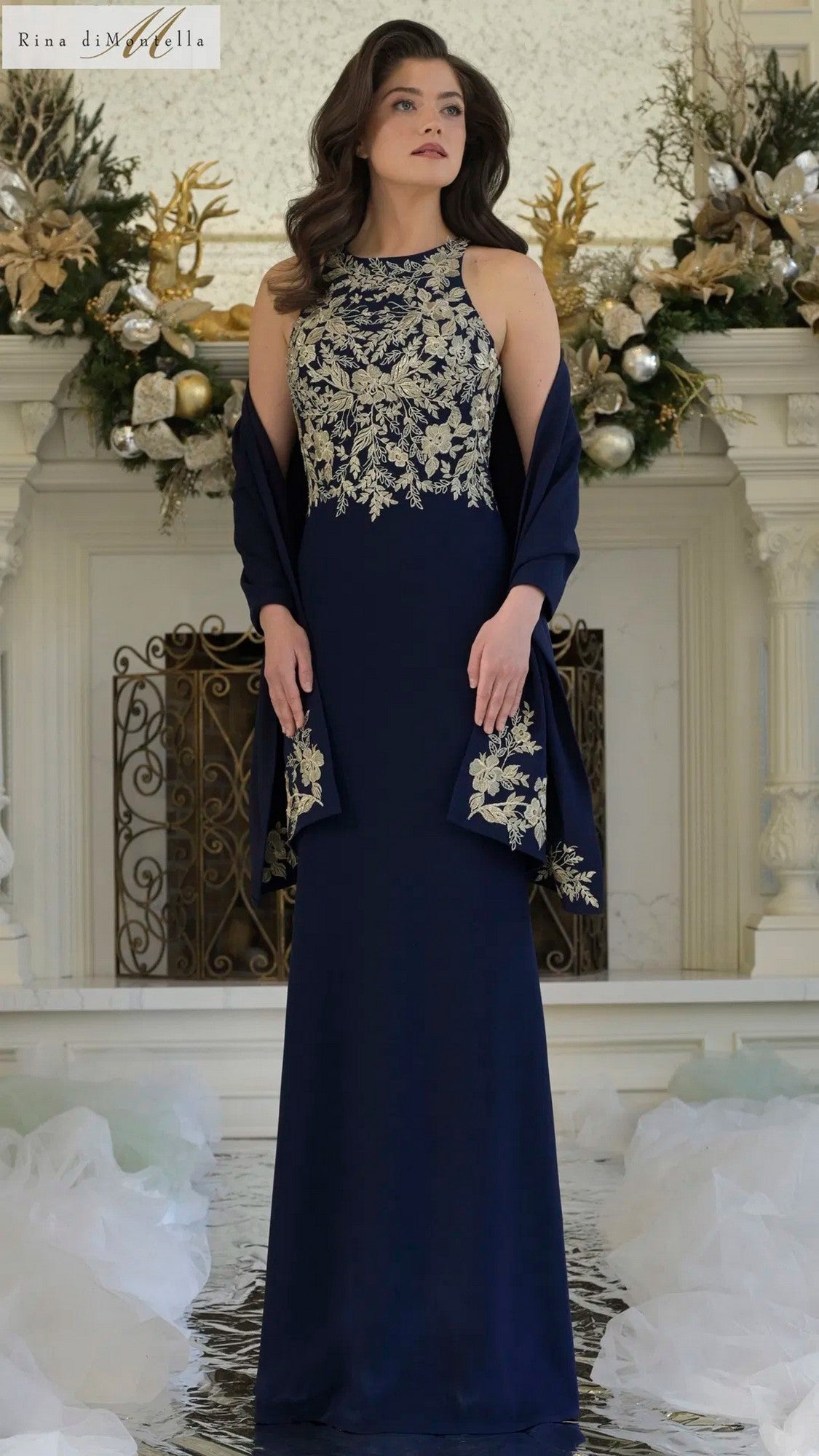 A woman stands indoors in a Rina Di Montella RD3345 long formal dress, navy gold with floral embroidery, holding a matching shawl. Festive decor accents the mantel behind her.